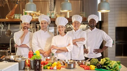 Multicultural work team of professional chefs is standing at workplace, looking at camera and posing. Group of male and female cooks takes foto together on background of restaurant kitchen.