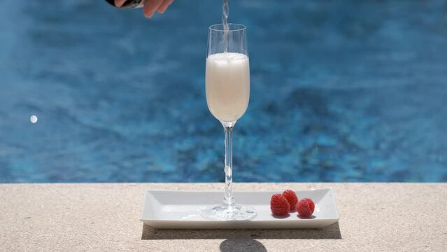 Close up of pouring glass with champagne or prosecco on swimming pool or sea background. Waiter pours cold champagne into glass. Sparkling white wine on tray with raspberry. Luxury alcohol drink. 4K