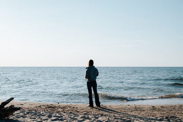 Lonely woman standing alone on seashore looking at seascape on sunny day, view from back