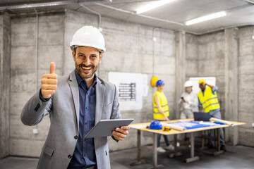Successful architect or project manager in business suit and hardhat holding thumbs up at construction site.