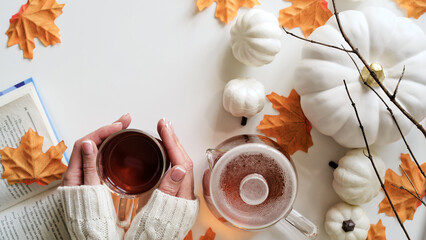 Female hands hold cup of tea,glass teapot,pumpkins,orange maple leaves,twigs and book stand nearby on white background,top view,flat lay,copy space.Concept of beginning of autumn,fall atmosphere,mood
