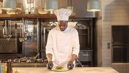 African American chef prepares puts plate with vegetable salad. Multicultural male chef dressed in special uniform cooking food in modern restaurant kitchen, looking at camera and smiling.