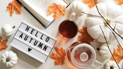 Close-up text board with inscription Hello Autumn,cup of tea, teapot,book,pumpkins,orange maple leaves,twigs on white background,top view,flat lay.Concept of beginning of autumn,fall atmosphere,mood
