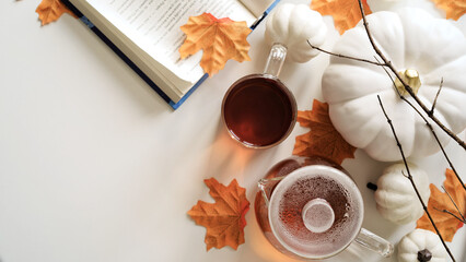 Close-up of cup of tea, glass teapot, pumpkins, orange maple leaves, twigs and book on white background, top view, flat lay, copy space. The concept of the beginning of autumn, fall atmosphere, mood.