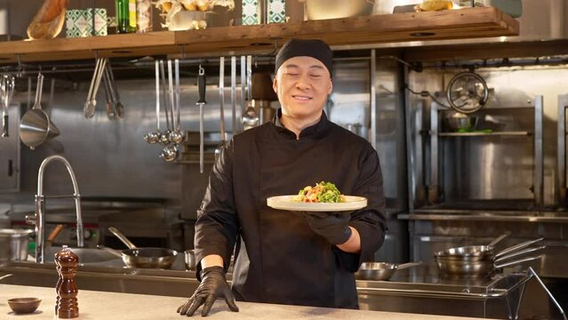 Asian man in special blac uniform stands in restaurant professional kitchen. Chef holding plate with vegetables salad, looking at camera and smiling on background of empty kitchen.