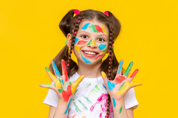 A young girl with brightly painted hands and multi colored pigtails shows her palms, smiling broadly. Yellow isolated background.