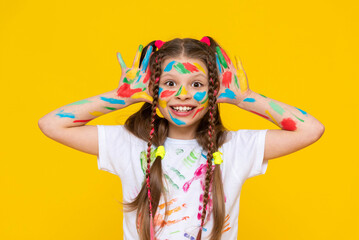 A young girl with brightly painted hands and multi colored pigtails shows her palms, smiling broadly. Yellow isolated background.