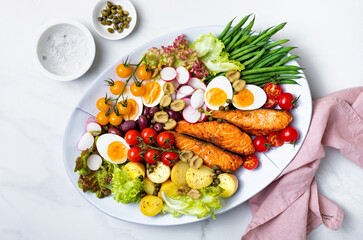 Delicious assorted grilled salmon nicoise salad with cherry tomatoes, eggs, olives and capers. Oval plate over white marble background