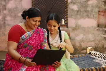 Happy Indian farmer mother and daughter using laptop sitting outside the home.