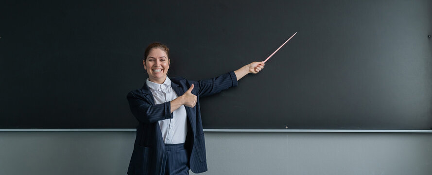 Red-haired caucasian woman in a pantsuit shows a thumbs up. Smiling female teacher with a pointer at the blackboard. Widescreen. 