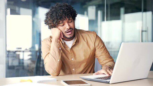 Overworked young employee is bored sitting at workplace at desk in modern office. Sleepy exhausted man in a shirt is tired from working with a laptop. A lazy male procrastinates, wants to sleep