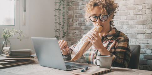 Modern woman at work online with laptop and wireless connection while eating a big sandwich for a fast lunch break too busy to relax. New business lifestyle freelance day. One female businesswoman
