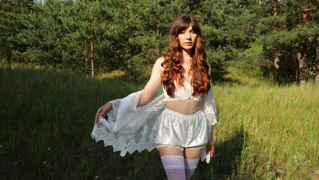 A beautiful young woman with long hair in white lingerie poses beautifully on the grass near the forest in the evening. Slow motion portrait woman brunette