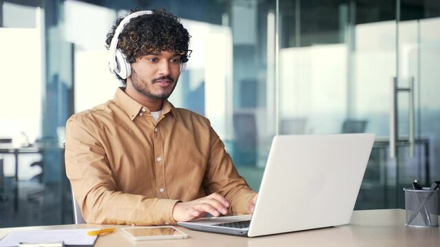 Young busy IT specialist coder in wireless headphones typing on laptop computer while sitting at workplace at desk in modern office. A handsome smiling developer in a shirt works on a project