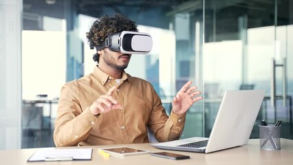 Young successful manager works using VR glasses in virtual reality simulator while sitting at workplace in modern office. Man uses gestures to control computer programs and flips through virtual pages - Powered by Adobe