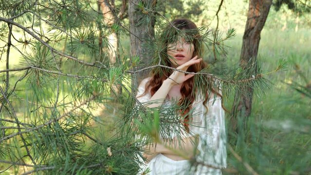 Portrait of a young brunette woman in white underwear hid behind pine needles in the forest. Slow motion erotica portrait