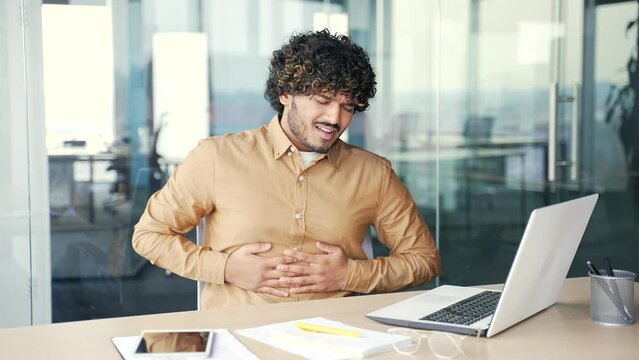 Young employee feels stomach pain while working on a laptop while sitting at workplace in modern office. Sick worker has heartburn, gastritis or poisoning. Male suffers from spasms and is constipated