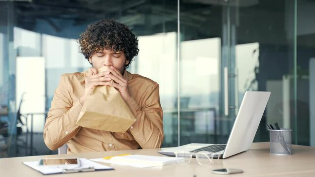 A young man having a panic attack while working on a laptop while sitting at a workplace at a desk in a modern office. The employee feels fear, breathes hard into a paper bag, is stressed and scared
