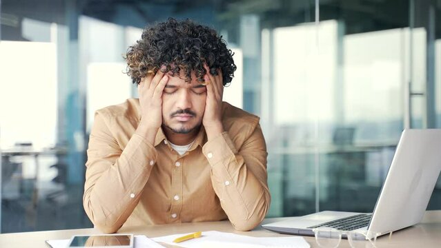 Young employee has depression suffering from mental pain sitting at workplace in modern office. Sad male holds his head with his hands, has personal problems, worries, feels despair and disappointment