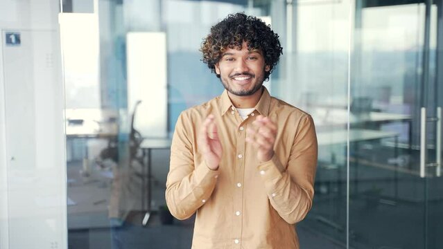 Portrait of a young smiling man applauding while standing in a modern office looking at the camera. Positive friendly businessman clapping hands expressing positive emotions and compliment performance