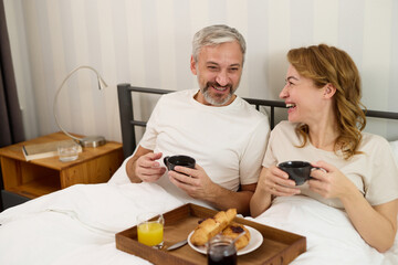 Mature couple enjoying coffee and breakfast at home in bed