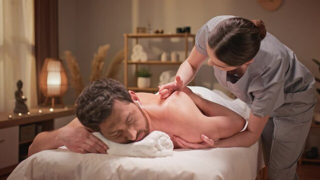 Attractive bearded man with earpods listening music during massage. Handsome patient resting while laying on bed in spa center. Young brunette massagist kneading man's back and stroking his skin.