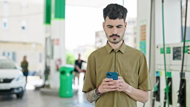 Young hispanic man using smartphone smiling at petrol station