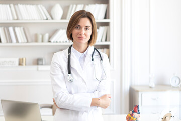 Portrait photo of young and beautiful smiling female doctor in white medical gown standing in the office of the modern clinic