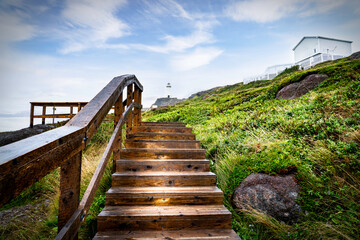 Wooden staircase with handrail leading up to a tall white lighthouse and historic buildings overlooking the Atlantic Ocean at Cape Spear Newfoundland Canada.