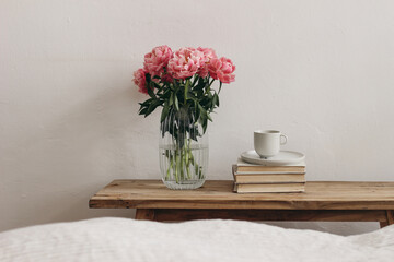 Feminine interior, still life. Rippled glass vase with coral pink peony flowers. Floral arrangement. Cup of coffee, books on vintage teak wooden bench, table. Empty white wall background. Blurred bed © tabitazn