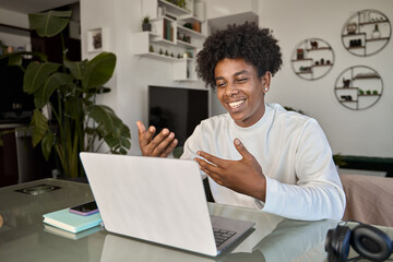 Happy African gen z teen student using laptop remote elearning at home table looking at computer...