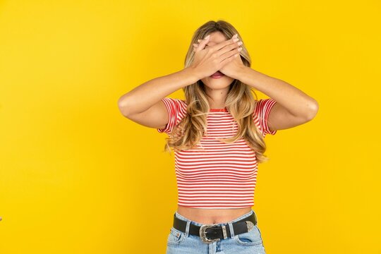 Young beautiful woman wearing striped t-shirt over yellow background Covering eyes and mouth with hands, surprised and shocked. Hiding emotions.
