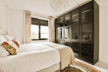 a bedroom with a white bed and black cabinetd wardrobes in the corner, next to a large window