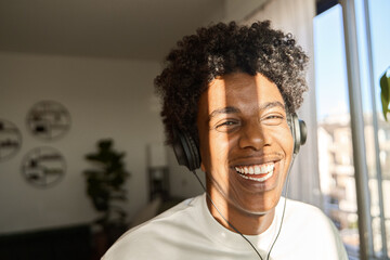 Happy funky gen z hipster African American guy wearing headphones laughing at home, listening...