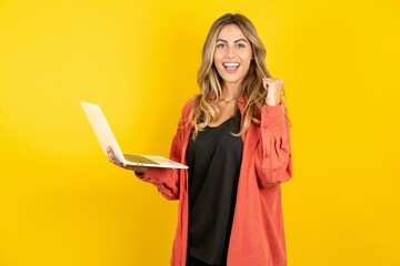 young beautiful blonde woman standing over yellow studio background hold computer open mouth rise...
