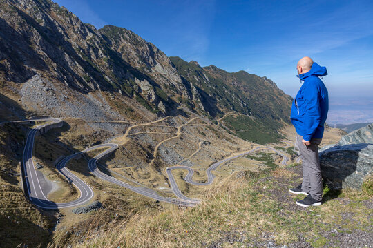 A tourist in a blue jacket looks at the road in the mountains, back view. Carpathians, Ridge Fagarash. Transfagarash road
