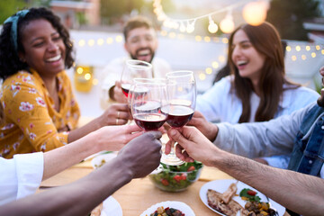 Smiling young people toasting red wine at rooftop barbecue. Gathered colleagues drinking and eating outdoors. Group of diverse friends enjoying free time on vacation. Relationships and celebrations.