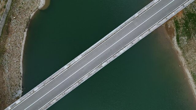 Aerial view of a bridge over a lake the road diagonally separates the image in half, a red car crosses the frame, the drone rises in altitude slightly widening the frame. 