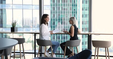 Two positive young and mature office colleagues women talking in office lobby, discussing project strategy, plan. Mature businesswoman speaking to coworker, teaching intern, new worker
