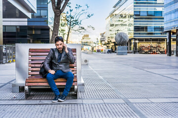 A businessman is talking on the cell phone while sitting on a bench in the street..