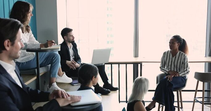 Positive confident young African expert woman giving consultation to diverse corporate team. Business leader asking questions from audience on training, giving educating lecture