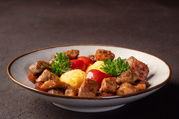 Romanian festive dish pomana porcului consisting of pieces of fried pork and sausages with pickled red peppers and polenta close-up on a plate on dark background. 

