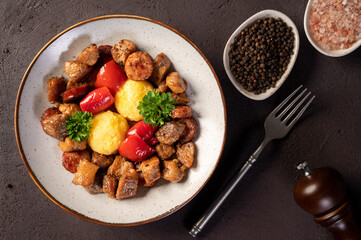 Romanian food pomana porcului made from chunks of fried pork and sausages served with corn porridge and pickled red peppers in a plate on a dark background. Horizontal Top View.
