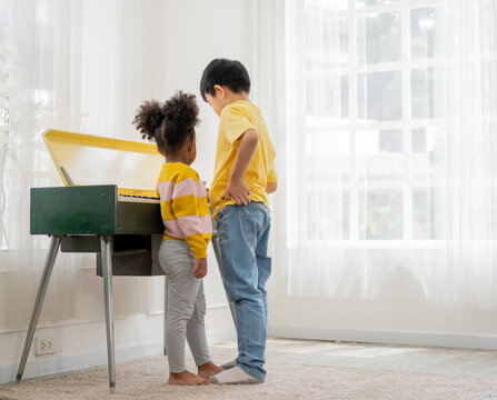 back view of boy and girl sibling standing playing piano together in living room apartment, diversity school children enjoy learning music lesson for creativity education in comfortable environment