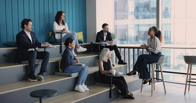 Young African American business leader woman holding speech, training diverse group of employees witting in rows on studying benches, giving lecture, workshop, seminar