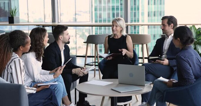 Positive confident mature business leader woman talking to team on corporate meeting in office lobby, speaking to diverse coworkers, smiling. Multiethnic group discussing teamwork strategy
