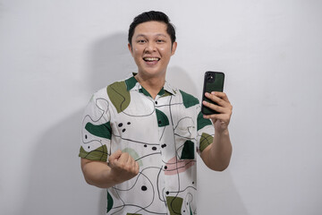 Young asian man wearing vintage shirt happily gesturing while looking at mobile phone, yes expression