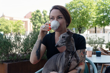 Young woman with her dog sitting in street friendly cafeteria and drink coffee.