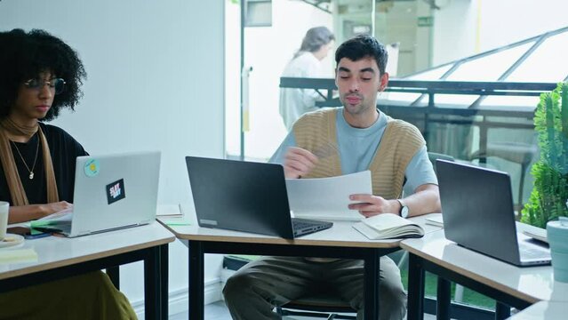 Young handsome gen Z businessman sitting at meeting table in modern coworking room, giving papers to multi-ethnic colleagues, holding team meeting