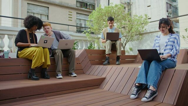 Group of gen Z business people in casual wear sitting on benches at public place in the city, using laptop and communicating, working remotely or studying online. Full shot